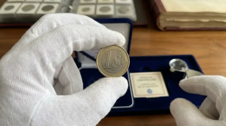 Mani con guanti bianchi mostrano una moneta da 1 euro, in un contesto da collezionismo numismatico