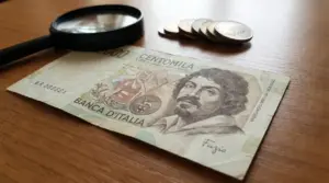 Banconota da 100.000 lire Caravaggio con firma Fazio su tavolo, accanto a una lente e alcune monete