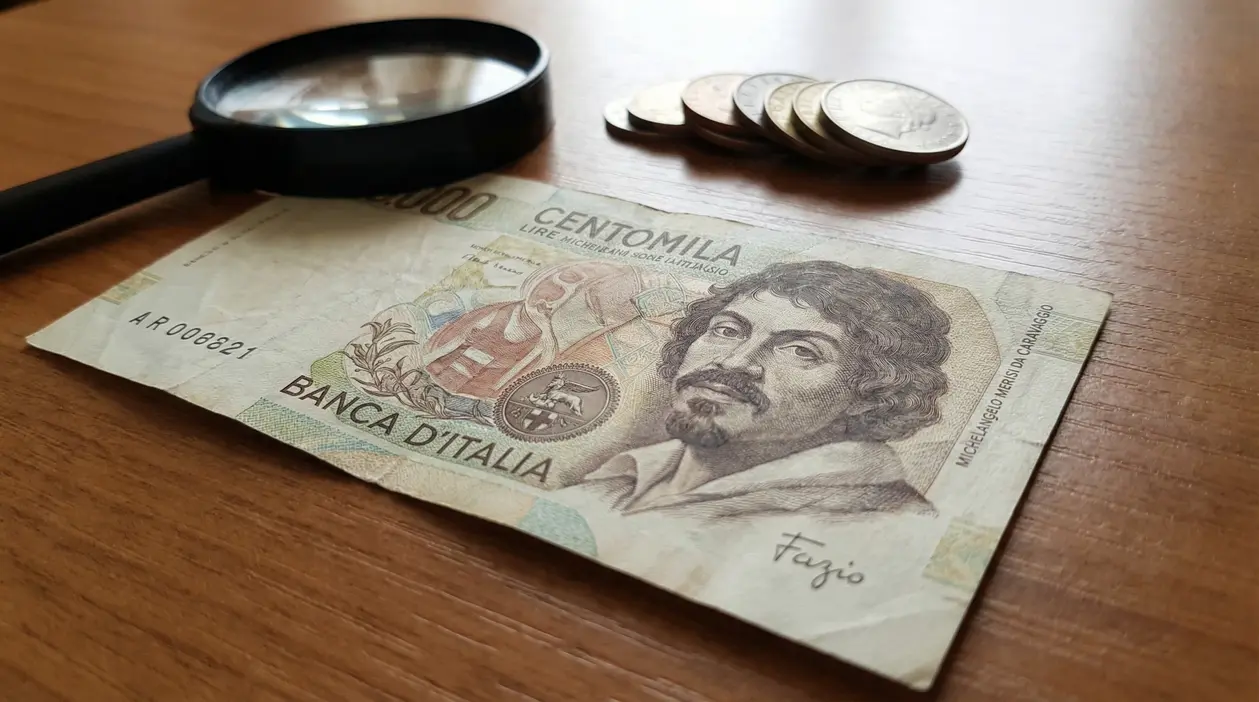Banconota da 100.000 lire Caravaggio con firma Fazio su tavolo, accanto a una lente e alcune monete