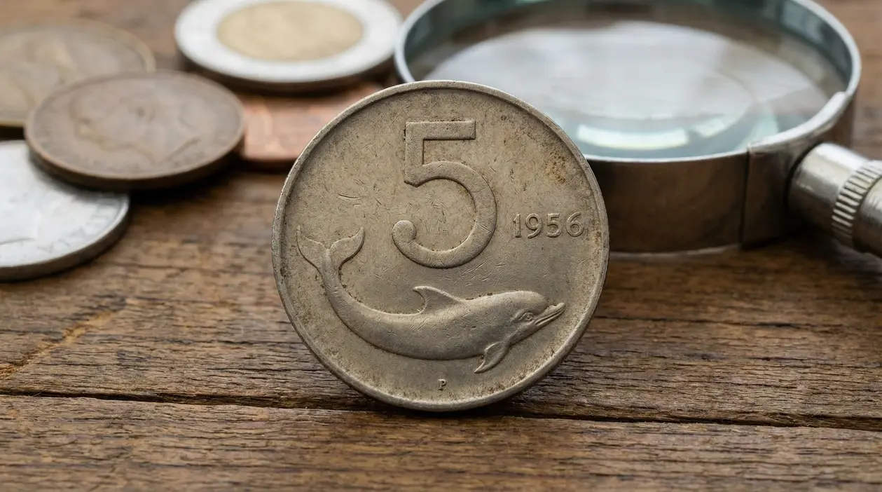 Moneta da 5 lire con delfino del 1956 in primo piano accanto a una lente su un tavolo di legno