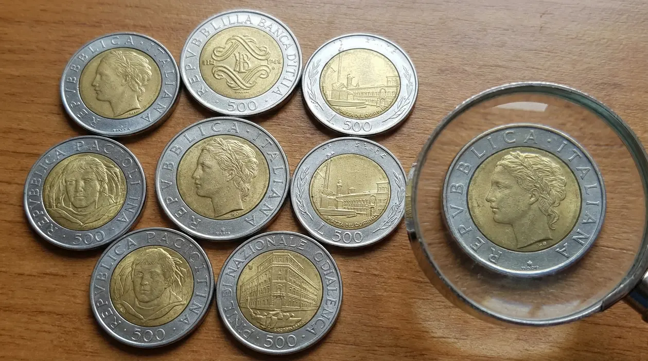 Monete italiane da 500 lire bimetalliche su tavolo in legno, con una osservata attraverso una lente