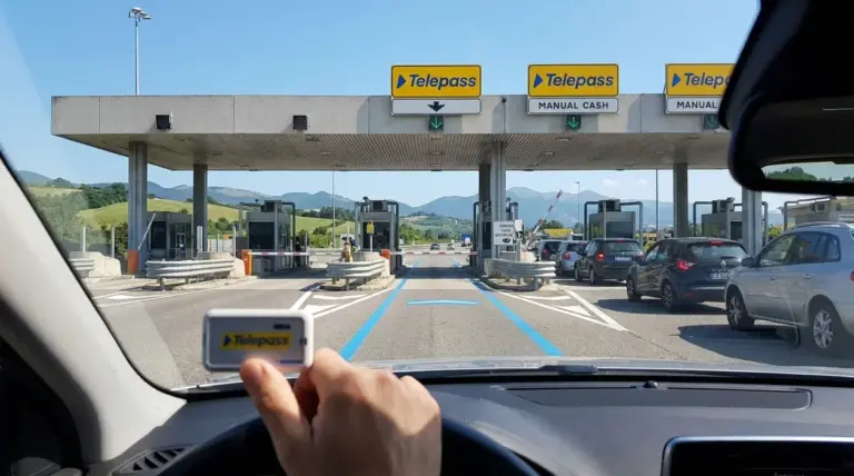 Casello autostradale con corsie di telepedaggio e auto in coda, visto dall’interno di un’auto con dispositivo in mano
