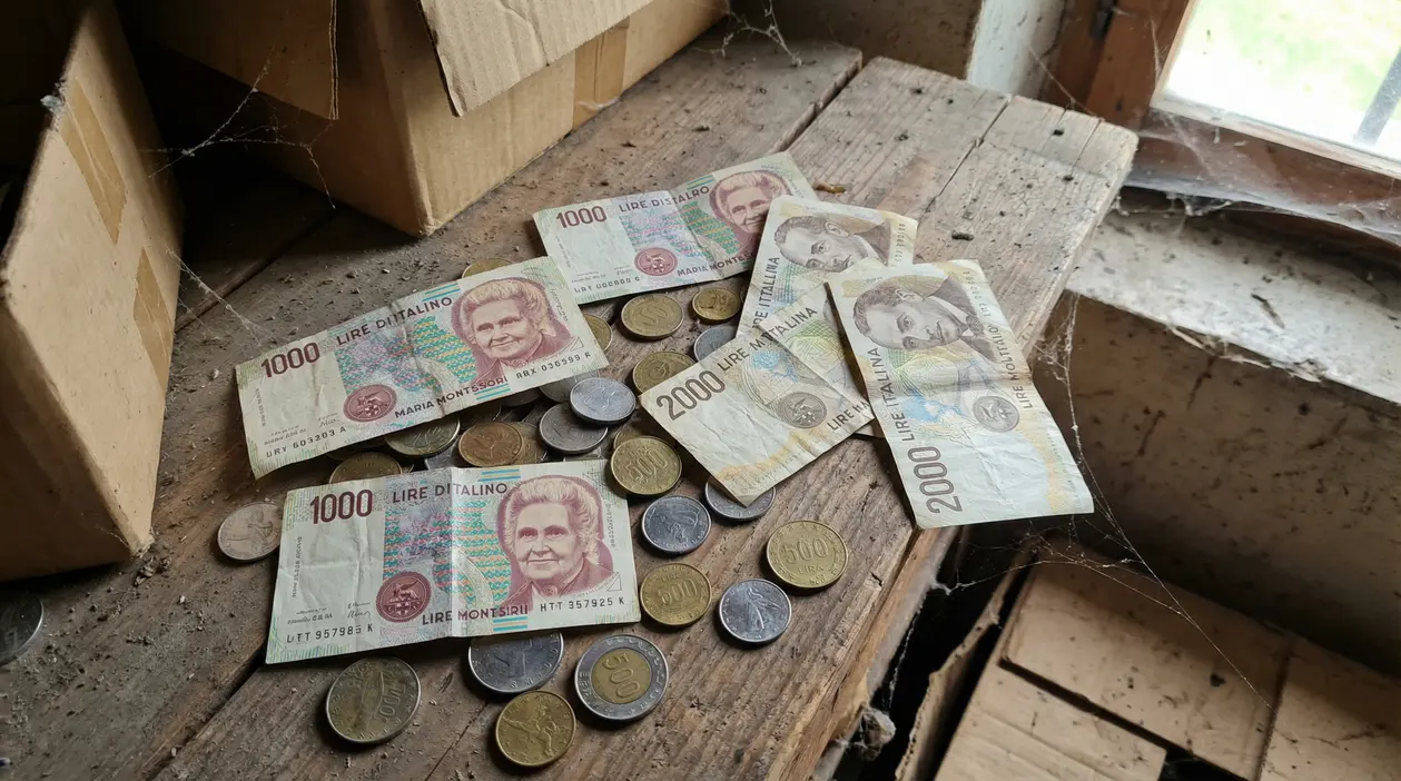 Vecchie banconote e monete in lire italiane su un tavolo polveroso accanto a scatole di cartone