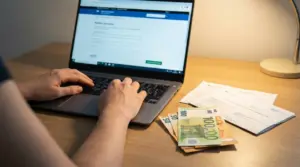 Persona che compila una domanda online su laptop, con banconote in euro e documenti sul tavolo