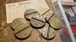 Gettoni telefonici vintage su un tavolo, accanto a una schedina del Totocalcio e a una cabina telefonica