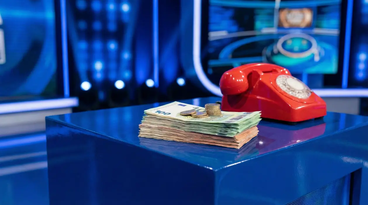Mazzetta di banconote e monete su un tavolo da studio TV, accanto a un telefono rosso, in un game show