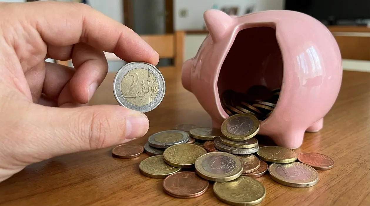 Mano che mostra una moneta da 2 euro accanto a un salvadanaio a forma di maialino rovesciato con monete sul tavolo