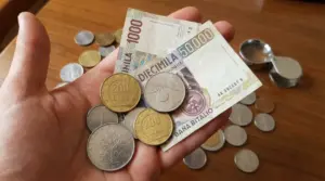 Mano che tiene banconote e monete in lire italiane su un tavolo di legno, con altre monete sullo sfondo