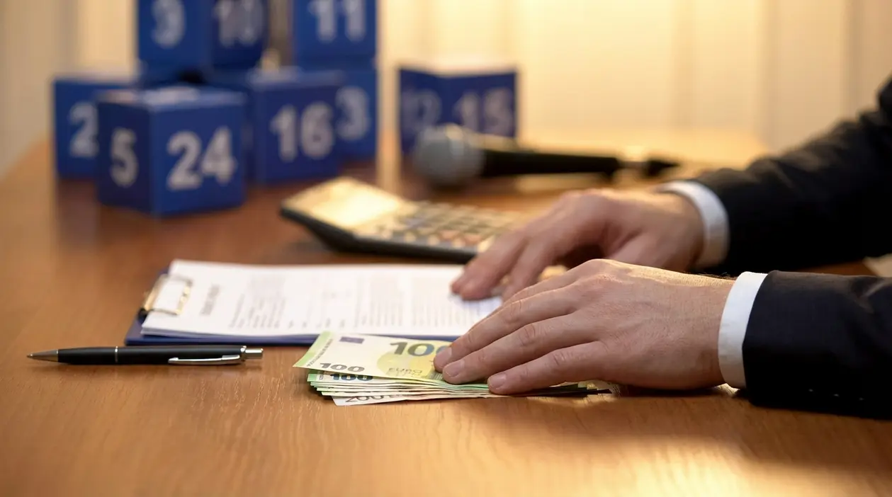 Mani su banconote in euro e documenti, con pacchi numerati sullo sfondo in un contesto legato ad Affari Tuoi