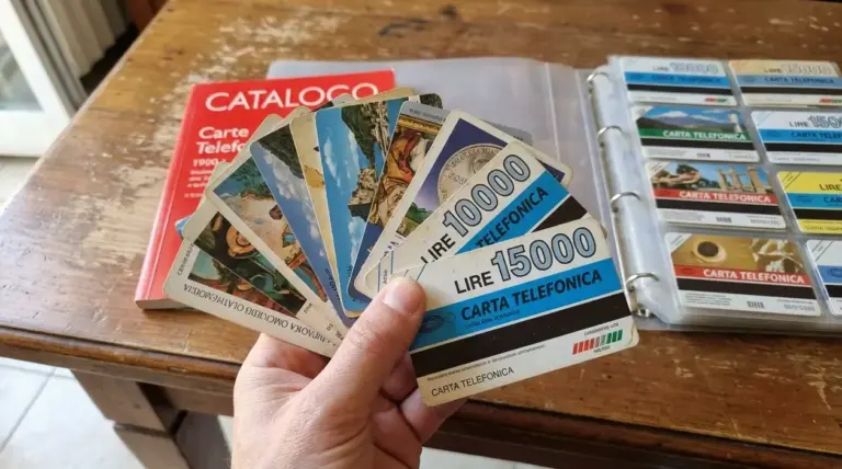 Mano che mostra vecchie carte telefoniche in lire accanto a catalogo e raccoglitore da collezione