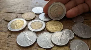 Monete da 50, 200 e 500 lire italiane sparse su un tavolo di legno, con una mano che ne mostra una