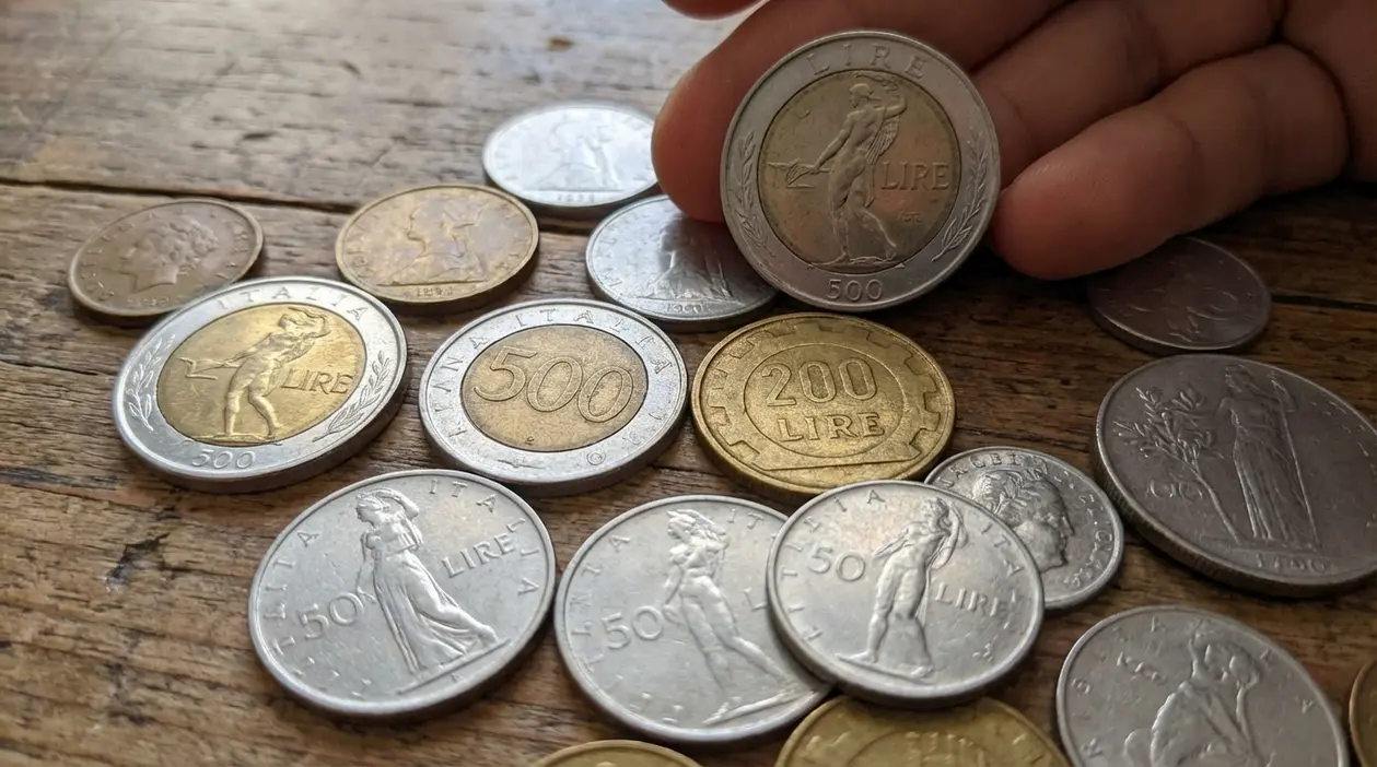 Monete da 50, 200 e 500 lire italiane sparse su un tavolo di legno, con una mano che ne mostra una